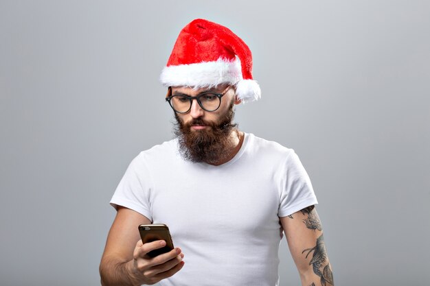 Weihnachten, Feiertage, Technologie und Menschenkonzept - gut aussehender bärtiger Mann in Weihnachtsmütze, der ein Selfie-Foto mit Smartphone auf grauem Hintergrund macht.