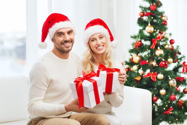 weihnachten, feiertage und menschenkonzept - glückliches paar in weihnachtsmützen mit geschenkboxen, die zu hause auf dem sofa sitzen