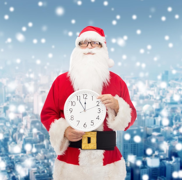 weihnachten, feiertage und personenkonzept - mann im kostüm des weihnachtsmanns mit uhr, die zwölf zeigende finger über schneebedecktem stadthintergrund zeigt