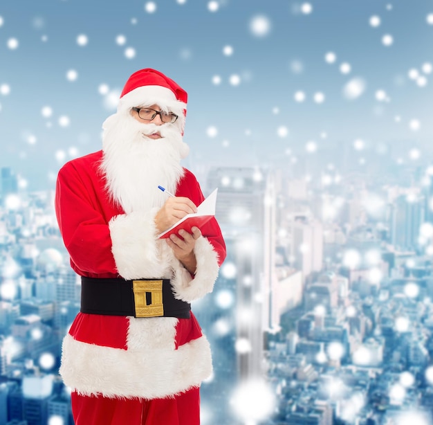 weihnachten, feiertage und personenkonzept - mann im weihnachtsmannkostüm mit notizblock und stift über schneebedecktem stadthintergrund