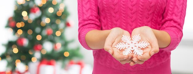 weihnachten, feiertage und personenkonzept - nahaufnahme einer frau in rosa pullover, die schneeflocke über wohnzimmer und baumhintergrund hält