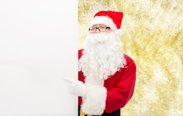 weihnachten, feiertage, werbung, gesten und menschenkonzept - mann im weihnachtsmannkostüm, der mit dem finger auf eine weiße leere plakatwand über gelbem lichthintergrund zeigt