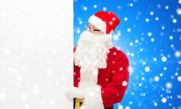 weihnachten, feiertage, werbung und personenkonzept - mann im weihnachtsmannkostüm mit weißer leerer plakatwand über blauem schneebedecktem hintergrund