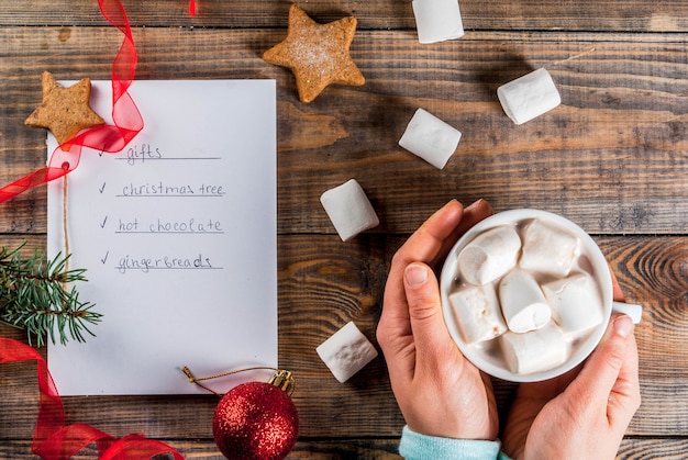 Weihnachten, Jamon. Holztisch, Notizbuch mit, zum der Liste zu tun, Mädchenhände hält Kakaobecher, Weihnachtsball, Kiefer, rotes Band, Eibisch. Ansicht von oben