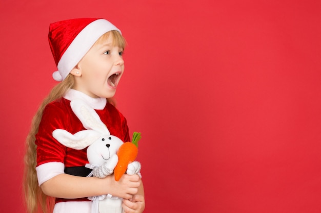 Weihnachten mit ihrer Freundin treffen. Kleines süßes Mädchen, das als Weihnachtsmann verkleidet wegschaut, schockiert mit offenem Mund hält ihr Spielzeugkaninchen