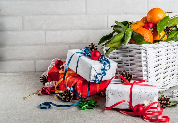 Weihnachten und. Frische Mandarinen mit grünen Blättern in einem weißen Korb, Weihnachtsdeko und Geschenkboxen, auf grauem Tisch,