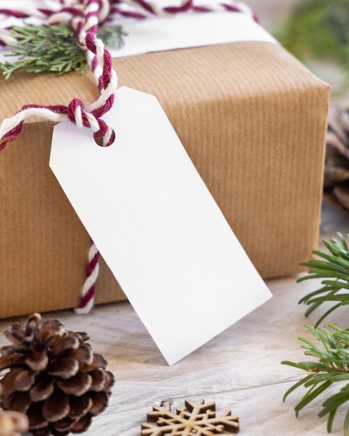 Weihnachten verpacktes Geschenk mit Papiergeschenkanhänger mit Tannenzweigen, Tannenzapfen und Feiertagsdekorationen aus nächster Nähe. Rustikale Winterkomposition mit leerem Geschenkanhänger Mockup, Kopierraum, Nahaufnahme