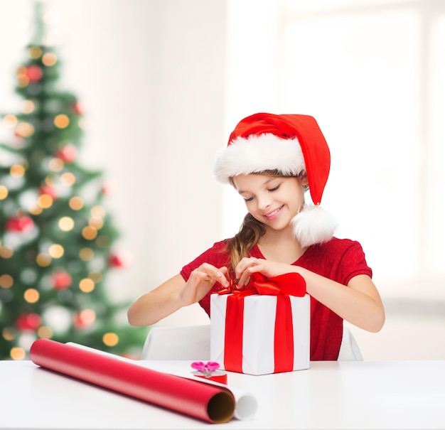 Weihnachten, Weihnachten, Winter, Glückskonzept - lächelndes Mädchen in Nikolausmütze mit Geschenkbox und Geschenkpapier