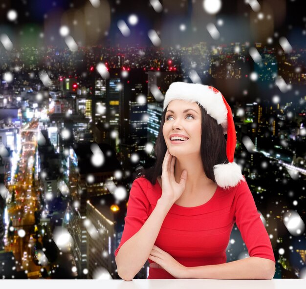 weihnachten, winter, feiertage, glück und personenkonzept - lächelnde frau in santa helfermütze über schneebedecktem nachtstadthintergrund
