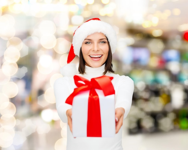 weihnachten, winter, glück, feiertage und personenkonzept - lächelnde frau in weihnachtsmannmütze mit geschenkbox über lichthintergrund