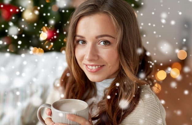 Weihnachten, Winterferien und Menschenkonzept - glückliche junge Frau mit einer Tasse Kaffee oder Kakao zu Hause über Schnee