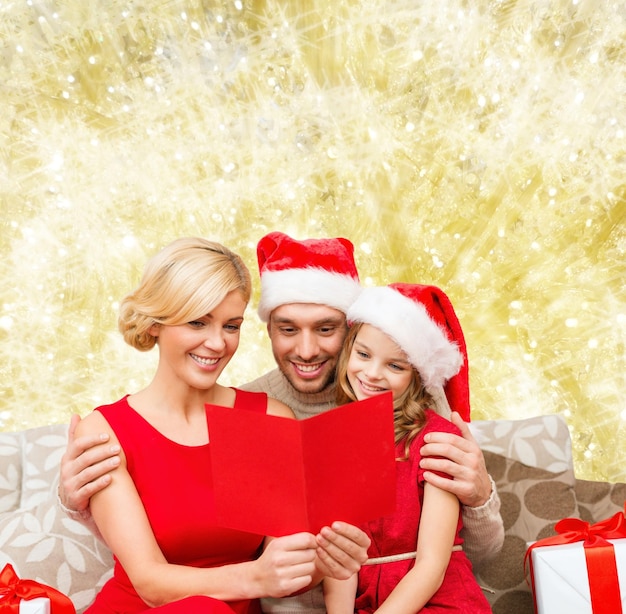 weihnachts-, feiertags-, familien- und menschenkonzept - glückliche mutter, vater und kleines mädchen in santa-helferhüten mit geschenkboxen, die geeting-karte über gelbem lichthintergrund lesen
