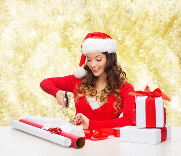 weihnachts-, feiertags-, feier-, dekorations- und menschenkonzept - lächelnde frau in weihnachtsmannmütze mit schere, die geschenkbox über gelbem lichthintergrund verpackt
