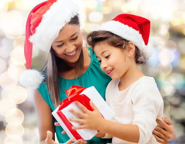 weihnachts-, feiertags-, feier-, familien- und personenkonzept - glückliche mutter und kleines mädchen in santa-helferhüten mit geschenkbox über lichthintergrund