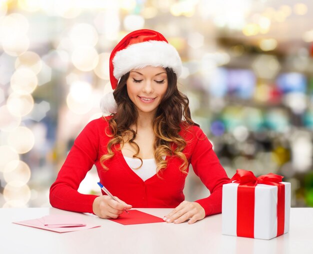 weihnachts-, feiertags-, feier-, gruß- und personenkonzept - lächelnde frau in weihnachtsmannmütze mit geschenkbox, die brief schreibt oder postkarte über lichthintergrund sendet
