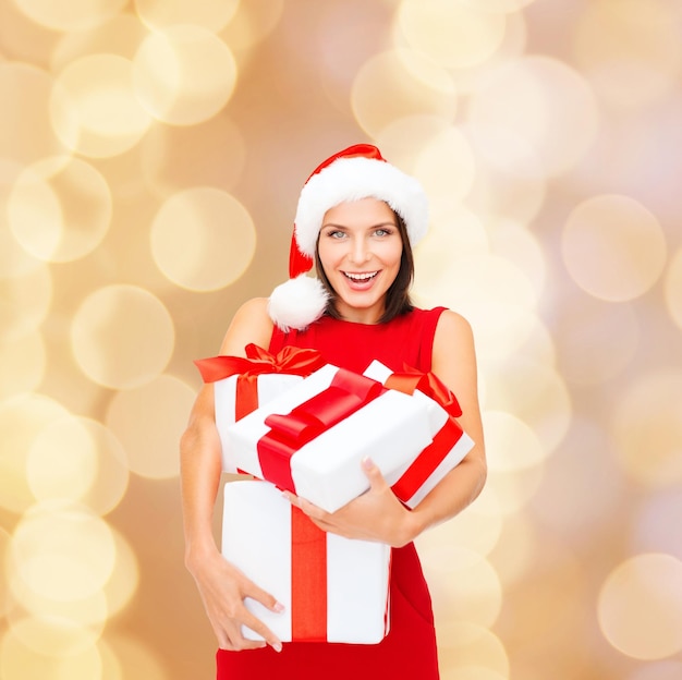 weihnachts-, feiertags-, feier- und menschenkonzept - lächelnde frau in weihnachtsmannmütze und rotem kleid mit geschenkboxen über beigem lichthintergrund