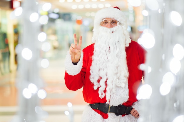 Weihnachts-, Feiertags-, Gesten- und Menschenkonzept - Mann im Kostüm des Weihnachtsmannes winkt mit der Hand über dem Hintergrund des Einkaufszentrums