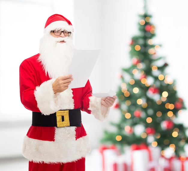 weihnachts-, feiertags- und personenkonzept - mann im kostüm des weihnachtsmannes, der brief über wohnzimmer mit baumhintergrund liest