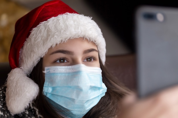 Weihnachts-Online-Grüße. Ein junges Mädchen in einem Weihnachtsmannhut und einer medizinischen Maske spricht unter Verwendung eines Smartphones für Videoanruffreunde und -eltern. Weihnachten während des Coronavirus