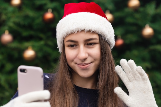 Weihnachts-Online-Grüße. Mädchen in Santa Claus Hut spricht und mit dem mobilen Smartphone für Videoanruf Freunde und Eltern. Weihnachten während des Coronavirus.