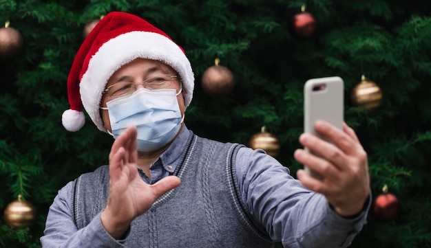 Weihnachts-Online-Grüße. Schließen Sie herauf Porträt des älteren Mannes, der einen Weihnachtsmannhut und eine medizinische Maske mit Emotion trägt. Vor dem Hintergrund eines Weihnachtsbaumes. Coronavirus Pandemie