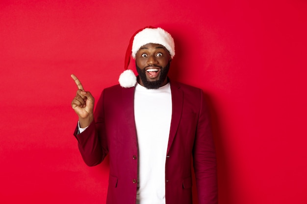 Weihnachts-, Party- und Feiertagskonzept. Aufgeregt und amüsiert Schwarzer Mann zeigt mit dem Finger nach links und lächelt, zeigt Logo und steht über rotem Hintergrund