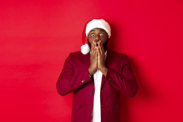 Weihnachts-, Party- und Feiertagskonzept. Schockierter Afroamerikaner in Weihnachtsmütze, der sprachlos auf Werbung reagiert und vor rotem Hintergrund steht.