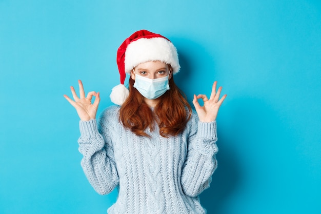 Weihnachts-, Quarantäne- und Covid-19-Konzept. Süßes rothaariges Teenie-Mädchen in Weihnachtsmütze und Pullover, das eine Gesichtsmaske aus Coronavirus trägt, in Ordnung zeigt, etwas genehmigt und lobt