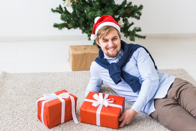 Weihnachts- und Feiertagskonzept - Glücklicher Mann in der Weihnachtsmütze, die ein Geschenk zu Hause im Wohnzimmer öffnet