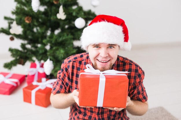 Weihnachts- und Feiertagskonzept - Lustiger Mann in der Weihnachtsmütze, die ein Geschenk zu Hause im Wohnzimmer hält