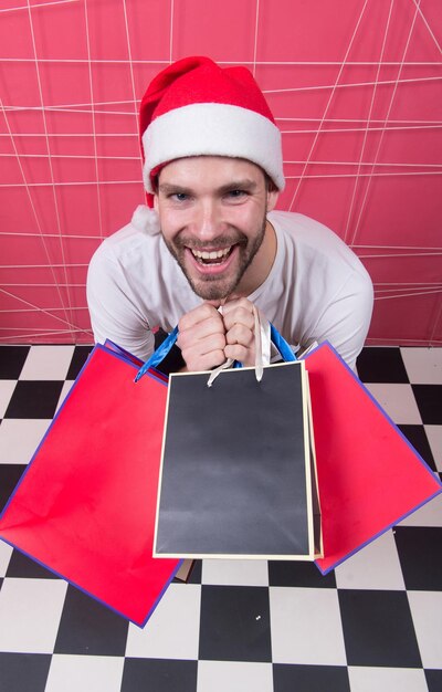 Weihnachts- und Neujahrsgeschenke. Mann im Weihnachtshutlächeln mit bunten Taschen. Winterschlussverkauf, Einkaufskonzept. Vorbereitung und Feier der Feiertage. Santa Shopper mit Paperbags auf rosa Hintergrund.