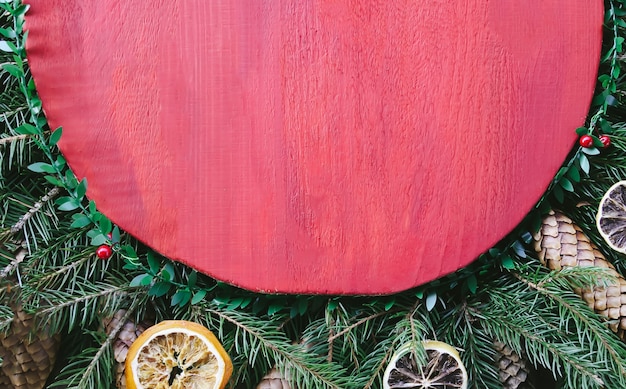 Weihnachts- und Neujahrswinterurlaubszusammensetzung mit roter Holzoberfläche auf grünen Tannenzweigen, getrockneten Orangenscheiben, Zapfen, Geschenkbox und roter Schleife.