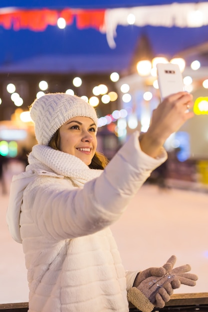 Weihnachts-, Winter-, Technologie- und Freizeitkonzept - glückliche junge Frau, die mit Smartphone auf der Eisbahn im Freien fotografiert.