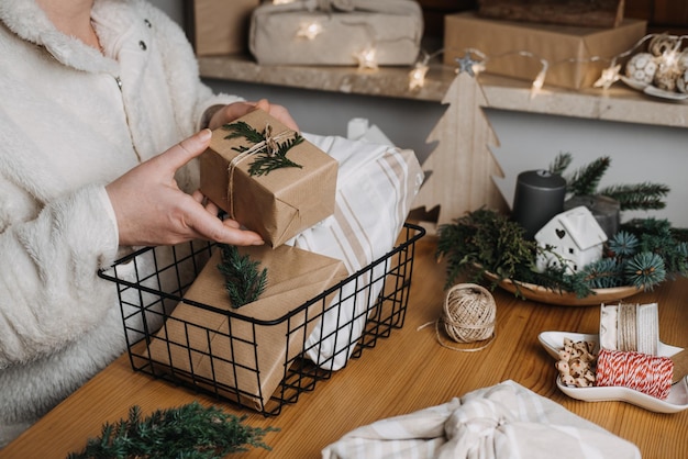 Weihnachts-Zero-Waste-umweltfreundlicher Wiederverwendungsprozess zum Verpacken von Geschenken Frau verpackt Weihnachts-Zero-Waste-wiederverwendbares Bastelpapier, nachhaltige Geschenke in Bastelpapier mit grünen Blättern auf dem Tisch zu Hause