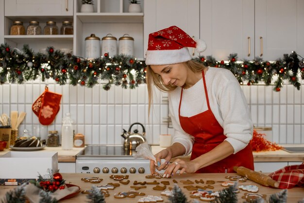 Weihnachtsbäckerei dekorieren. Frau mit Weihnachtsmütze, die hausgemachte Lebkuchen auf dem Küchentisch verglast