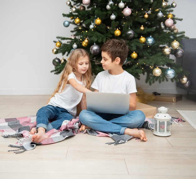 Weihnachtsbaum in einem großen Wohnzimmer. Kinder spielen in der Nähe des Weihnachtsbaums. Ein Junge mit einem Laptop kommuniziert mit Freunden.