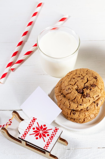 Weihnachtsbild mit Glas Milch gestreifte Strohhalme Cookies leere Notiz dekorativen Schlitten auf weißem Tisch