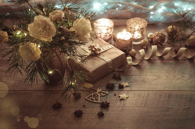 Weihnachtsdekoration. Geschenk, Kerzen, Lichter, goldene Kugeln auf einem rustikalen Holztisch. Weihnachtskomposition aus Tannenzweigen und englischen Rosen in einer Vase. Goldene und bräunliche Ästhetik.