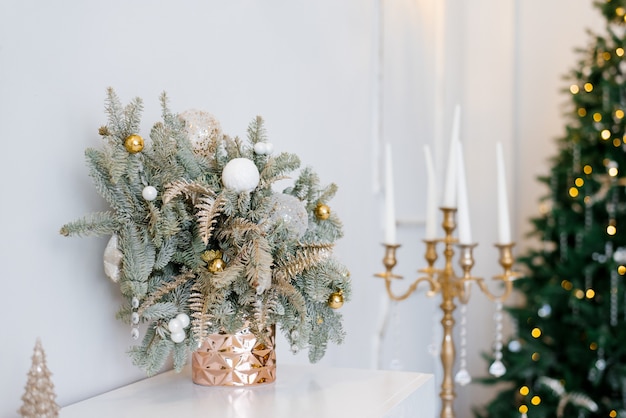 Weihnachtsdekoration in einem klassischen Wohn- oder Schlafzimmer in hellen Farben. Fichtenzweige in goldenen Vasen mit Spielzeug und einem goldenen Kerzenhalter auf der Kommode