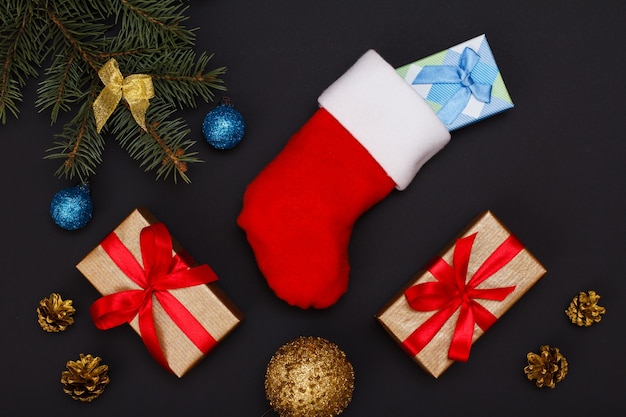 Foto weihnachtsdekoration. santa's boot, geschenkboxen, tannenbaumzweige mit zapfen und weihnachtsspielzeug auf schwarzem hintergrund. ansicht von oben. weihnachtsgrußkartenkonzept.