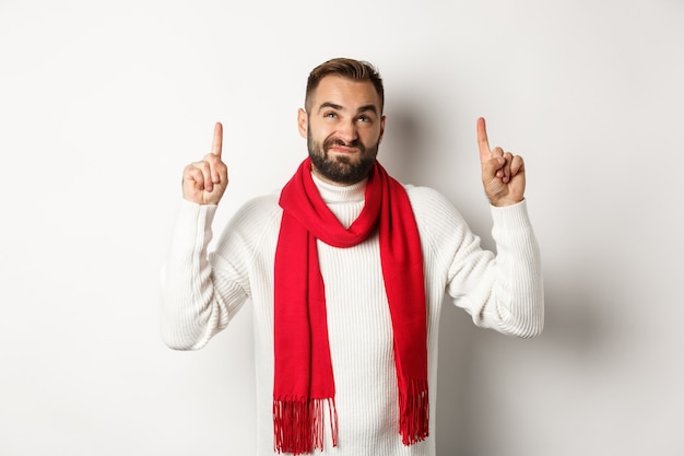 Weihnachtseinkaufs- und Winterferienkonzept. Unzufriedener bärtiger Kerl, der sich beschwert, mit dem Finger nach oben zeigt und zögerlich aussieht und auf weißem Hintergrund steht