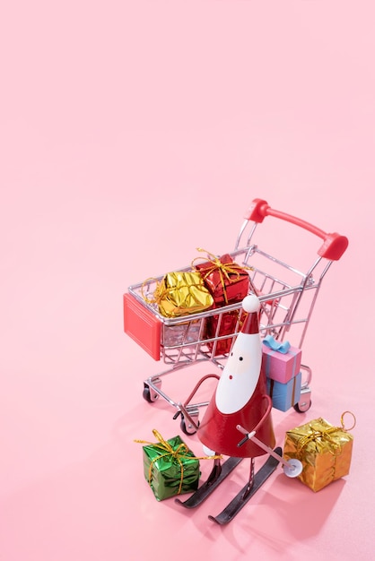 Weihnachtseinkaufskonzept Mini-roter Einkaufswagenwagen mit Weihnachtsmann-Spielzeug und Geschenkbox isoliert auf blassrosa Hintergrund Leerer Kopierraum Nahaufnahme