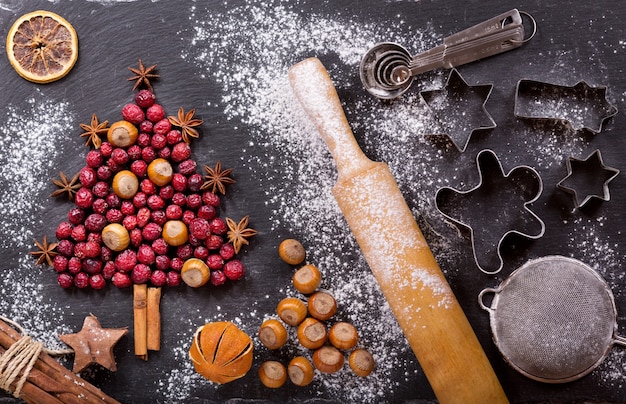 Weihnachtsessen. Zutaten zum Kochen von Weihnachtsbäckerei: Tannenbaum aus getrockneten Preiselbeeren mit Nüssen, Küchenutensilien und Trockenfrüchten auf einem dunklen Tisch, Draufsicht