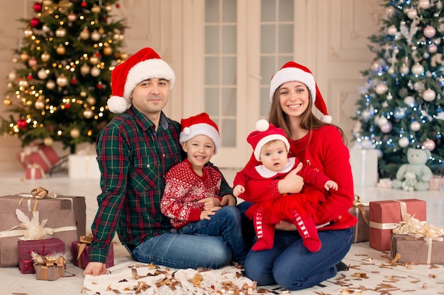 Weihnachtsfamilie! Glückliche Mutter, Vater und kleine Tochter und Sohn auf Weihnachtsmannmütze. Liebe Umarmungen genießen, Urlaub Menschen. Weihnachtsbaum mit Girlanden im verzierten Raumhintergrund. Zusammengehörigkeitskonzept