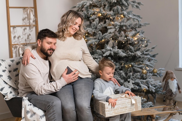 Weihnachtsfamilienglück Porträt von Papa, schwangerer Mutter und kleinem Sohn, der zu Hause im Sessel in der Nähe des Weihnachtsbaums sitzt, umarmt das Lächeln des europäischen jungen Erwachsenenfamilienurlaubsmorgens