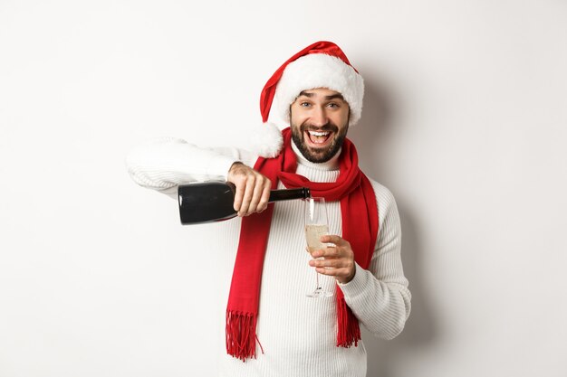 Weihnachtsfeier und Feiertagskonzept. Glücklicher bärtiger Mann in Weihnachtsmütze und Scard, gießt sich Champagner ein und lächelt, feiert Neujahr, steht auf weißem Hintergrund