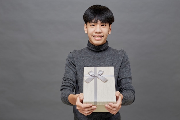 Weihnachtsfeier und Feiertagskonzept. junger asiatischer Mann mit Geschenkbox Studioaufnahme, auf grauem Hintergrund isoliert