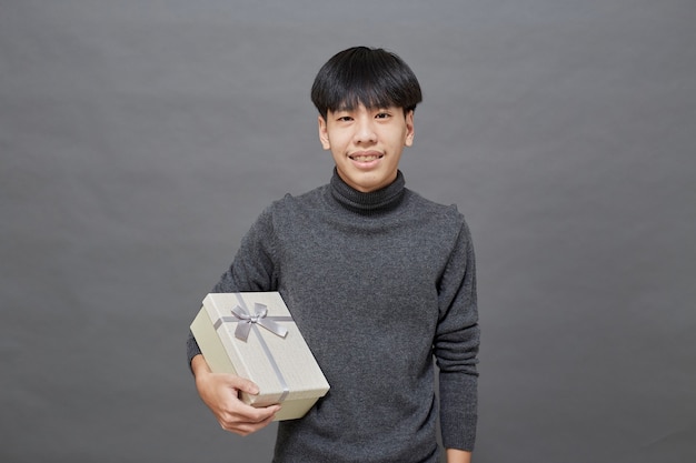 Weihnachtsfeier und Feiertagskonzept. junger asiatischer Mann mit Geschenkbox Studioaufnahme, auf grauem Hintergrund isoliert