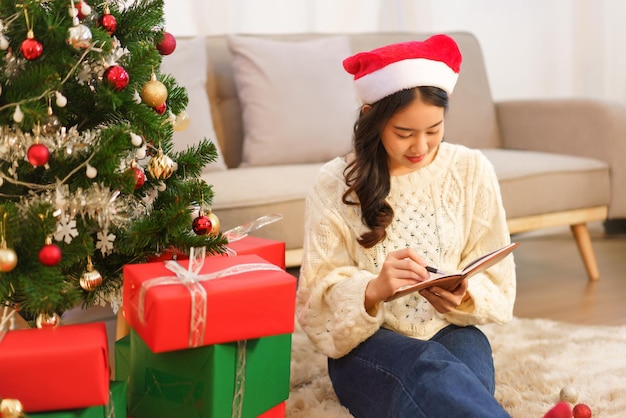 Weihnachtsfeierkonzept Asiatische Frau sitzt auf dem Boden in der Nähe des Weihnachtsbaums, um in ein Notizbuch zu schreiben