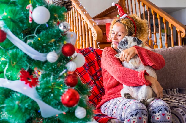 Weihnachtsfeiertag zu Hause mit einer glücklichen kaukasischen jungen Frau und einem lustigen süßen, entzückenden Mopshund, der sich umarmt und zusammen Spaß hat